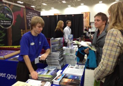 A man stands behind a table at a booth while talking to people.