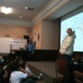 A man speaks to a group of students with a map in the background.