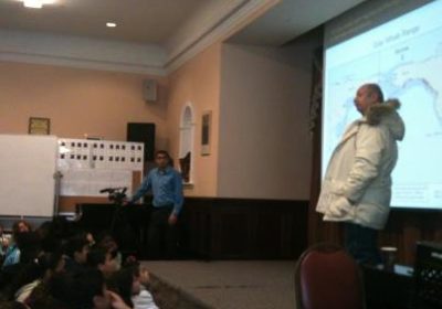 A man speaks to a group of students with a map in the background.