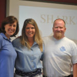 Two women and a man stand in front of a sharks sign.