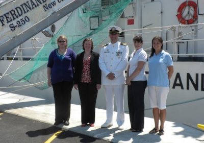Four women stand around a man in a military uniform in front of a ship.