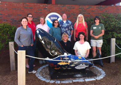 Nine people stand outside in front of a NOAA sign and behind an art sculpture of a whale