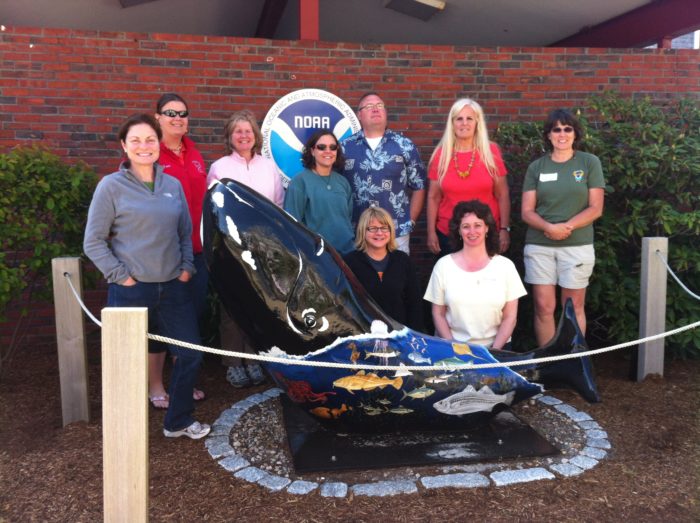 Nine people stand outside in front of a NOAA sign and behind an art sculpture of a whale