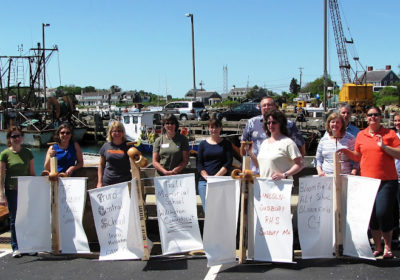 Eleven people stand holding drifter buoys with ships in the background.