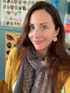 A headshot of woman with long, brown hair wearing a yellow sweater with a scarf and standing in a classroom.