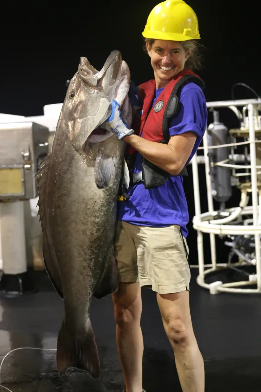 A woman in a hard hat standing on the deck of a ship holding a grouper that is almost as long as she is tall.