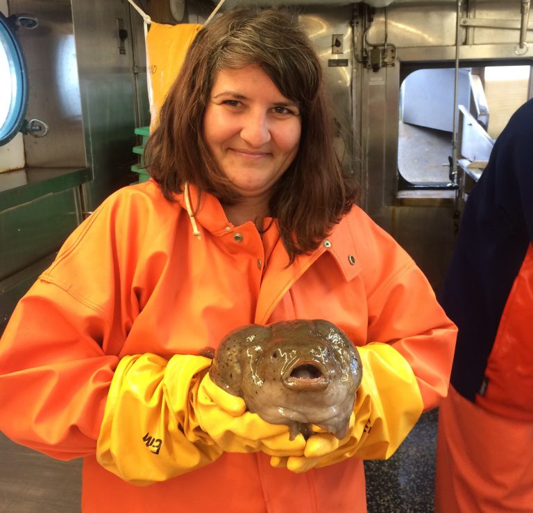 A woman with brown hair, wearing an orange rain slicker and yellow gloves. Standing in a ship's laboratory and holding a lumpsucker fish.