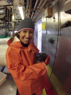 A woman in a ship's laboratory in a knit hat and orange rain slicker and wearing orange rubber gloves holding a lumpsucker fish.