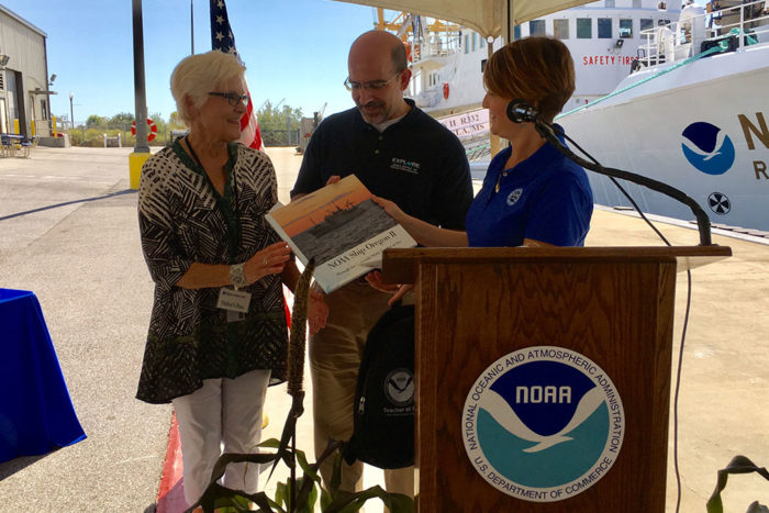 Two women and a man are outside and are standing in front of a podium with a NOAA emblem on it. All are standing in front of a NOAA ship. One of the women is handing an award to the man.