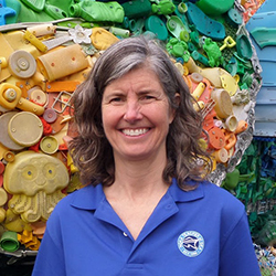 A woman in a blue Teacher at Sea shirt posing in front of marine debris art.