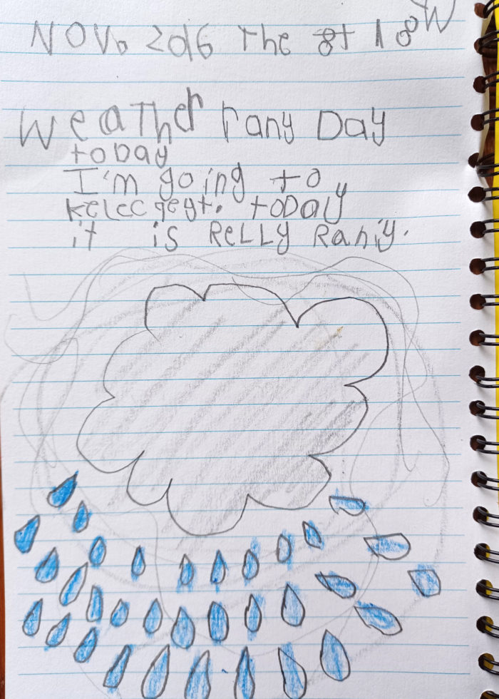 A pencil and colored pencil drawing by a young child of a cloud with rain drops falling from, done on notebook paper. The caption on the paper describes a rainy day.
