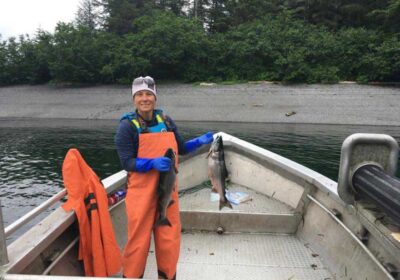 a fisherwoman on a small boat heading towards a sandy beach is holding a fish in each hand