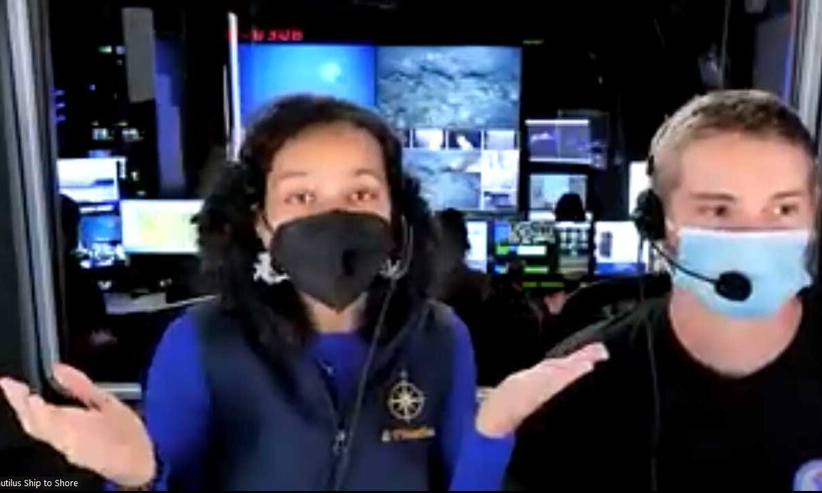 Two people wearing face masks in a control room with multiple screens displaying underwater images. the woman in a blue jacket gestures animatedly, the man, also in a mask, listens.