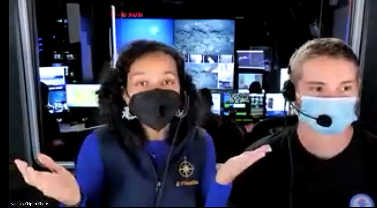 Two people wearing face masks in a control room with multiple screens displaying underwater images. the woman in a blue jacket gestures animatedly, the man, also in a mask, listens.