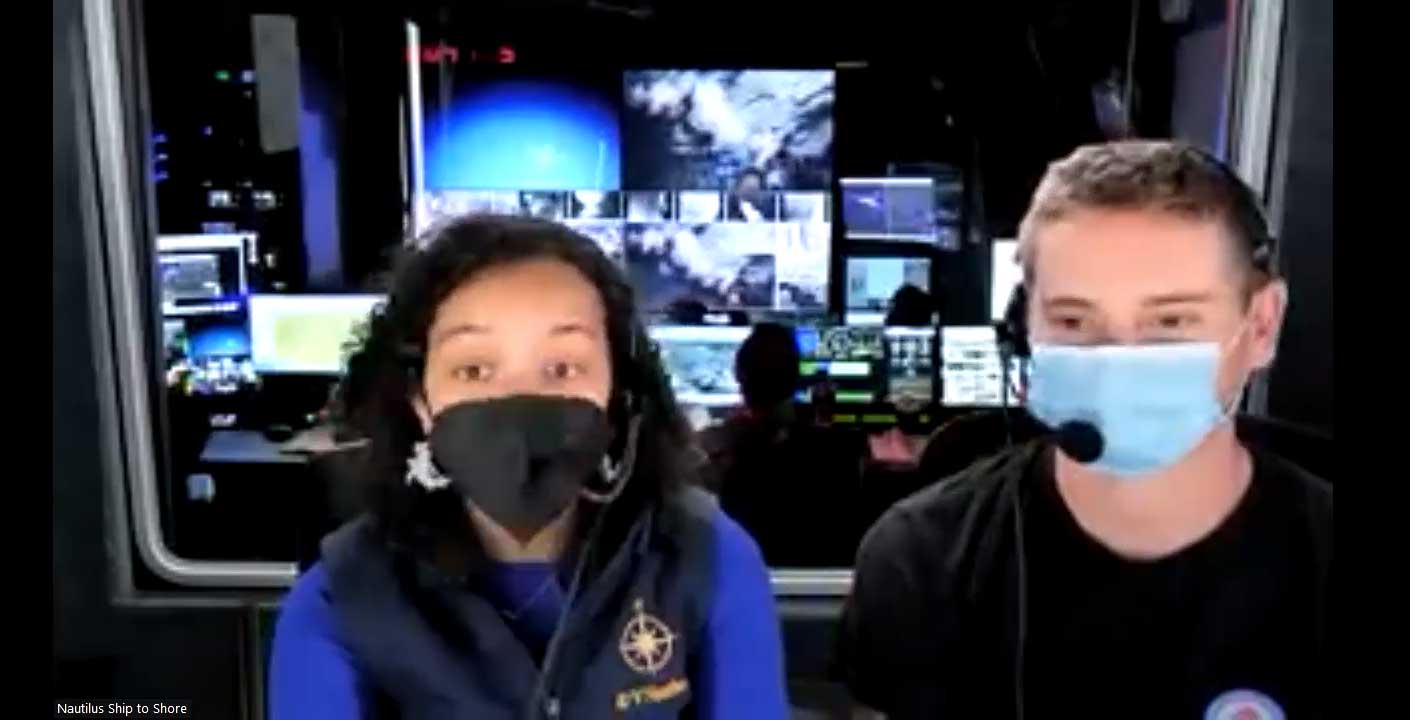 Two people, one wearing a black mask and one wearing a blue mask, sitting in a ship-to-shore control room filled with multiple screens displaying various data.