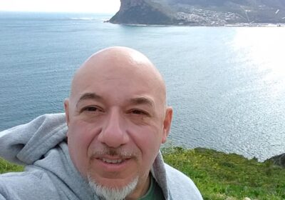 A man with a shaved head and a smile, wearing a hoodie, stands in front of a coastal landscape with sharp cliffs and the ocean behind him.