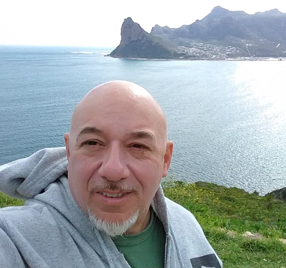 A man with a shaved head and a smile, wearing a hoodie, stands in front of a coastal landscape with sharp cliffs and the ocean behind him.