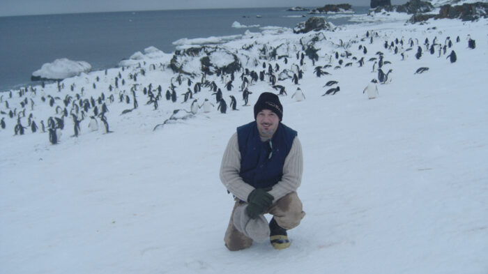 A man in winter clothing sits on snow-covered ground, smiling at the camera, with numerous penguins in the background on a rocky, snow-covered coastline.