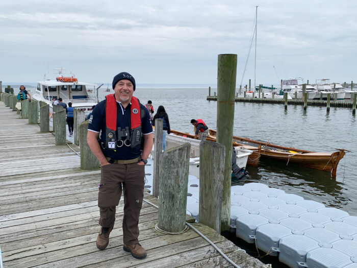 A man in a life jacket standing on a wooden dock, smiling with boats and the sea in the background on a cloudy day.