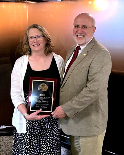 A woman and a man smiling as they hold a plaque together. the woman wears glasses and a polka dot skirt; the man sports a suit and tie. the plaque reads "bge safety leadership award.