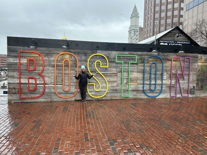 A person standing in front of a large, colorful "boston" sign with multicolored letters on a rainy day, wet pavement, and a building in the background.