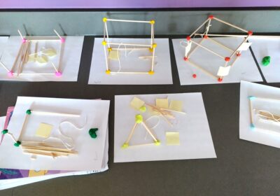 Multiple student projects on a table, featuring geometric structures made from sticks and connectors like clay, some with prominent yellow and red nodes, displayed on white sheets of paper.
