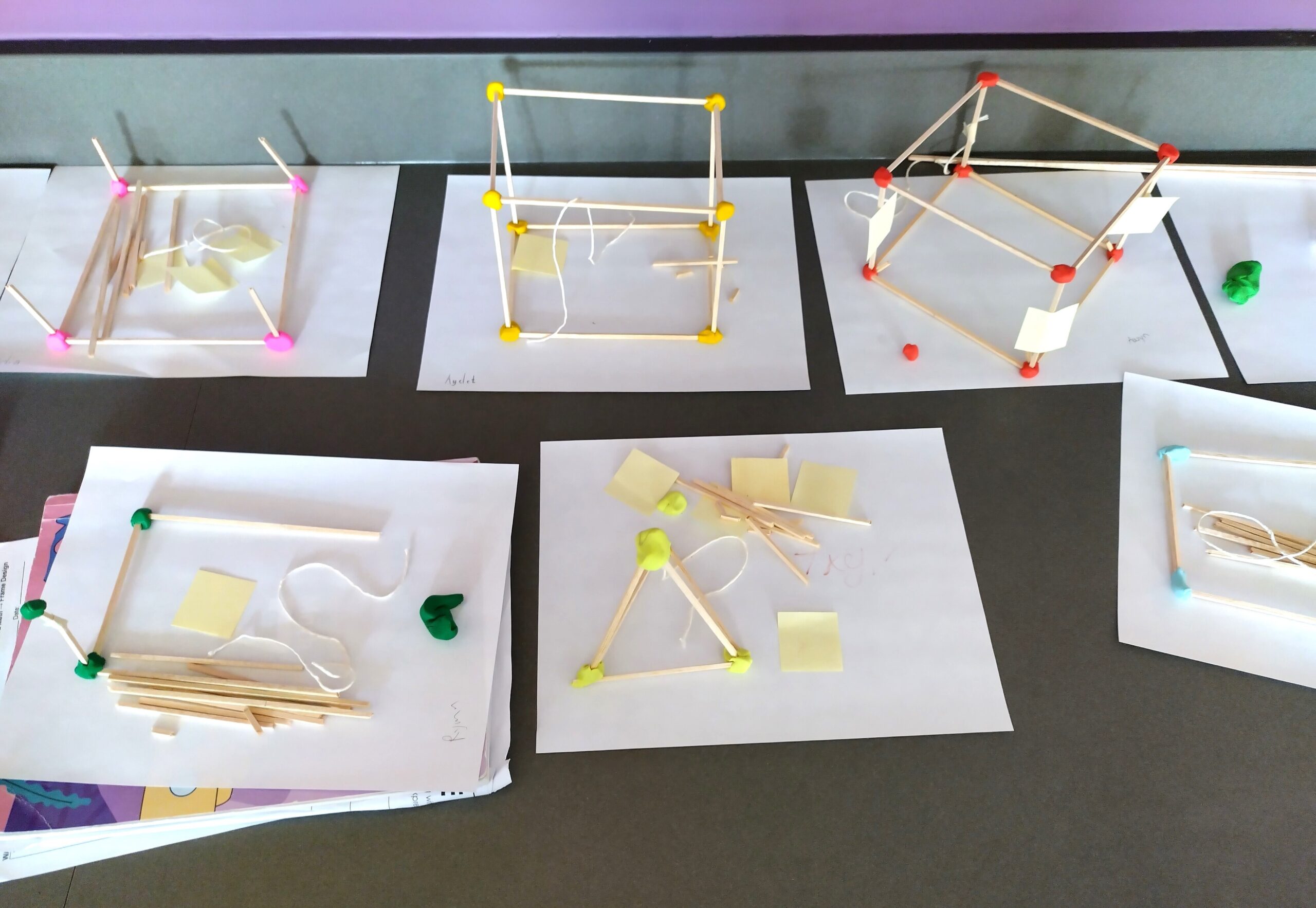 Multiple student projects on a table, featuring geometric structures made from sticks and connectors like clay, some with prominent yellow and red nodes, displayed on white sheets of paper.