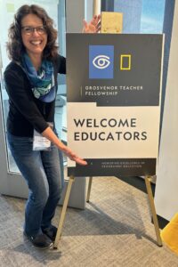 A joyful woman with glasses and a scarf poses beside a sign reading "Grosvenor Teacher Fellowship" and "Welcome Educators," promoting excellence in geographic education.