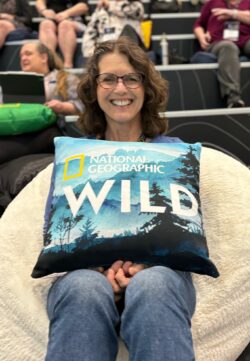 A smiling woman with glasses holds a "National Geographic WILD" pillow in a stadium setting with people in the background.