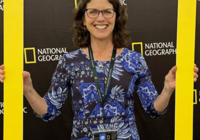 A woman in a blue patterned dress and glasses smiles while holding a large National Geographic frame. She wears a badge with the name "Jenny Heritage.