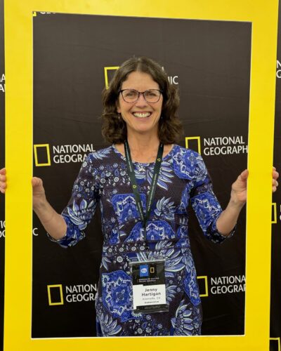 A woman in a blue patterned dress and glasses smiles while holding a large National Geographic frame. She wears a badge with the name "Jenny Heritage.
