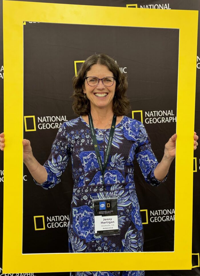 A woman in a blue patterned dress and glasses smiles while holding a large National Geographic frame. She wears a badge with the name 
