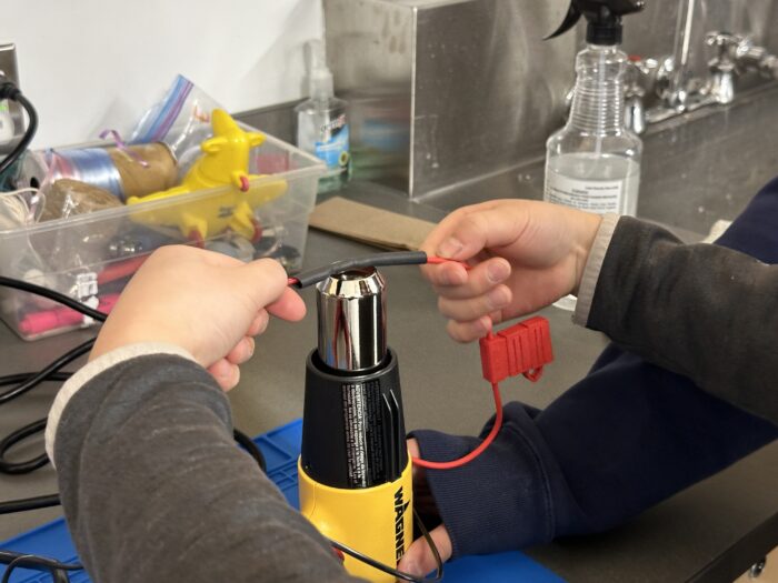 Two children using a yellow heat gun to perform a craft or technical task in a workshop setting. They are handling a black cable and a red component near the heat gun. A plastic container with small objects and a spray bottle are visible in the background.