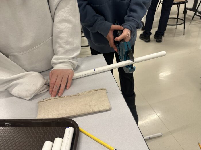 Two individuals work on a project at a table. One person holds a white PVC pipe on a cloth while the other uses a power tool to cut it. Other tools and materials, including a pencil and a tray, are on the table.