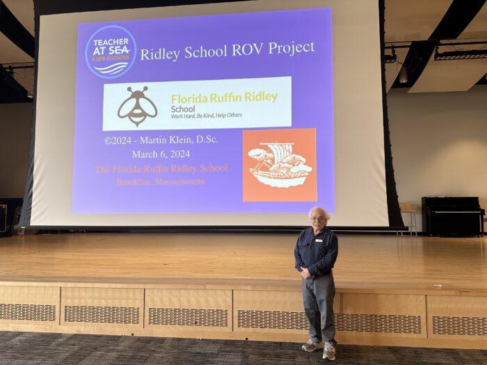 An older man stands in front of a large projection screen on a stage. The screen displays the title "Ridley School ROV Project" and includes logos and text about the project and a school. The man is wearing a dark long-sleeve shirt and jeans.