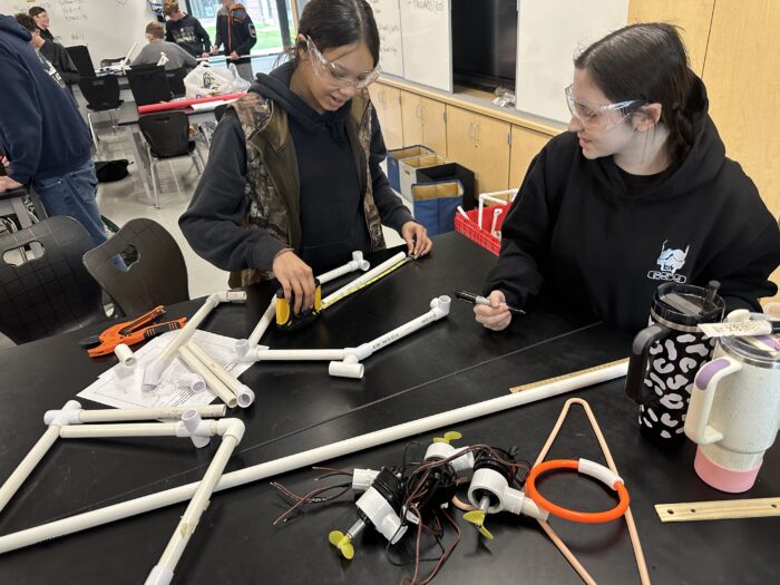 Two students wearing protective eyewear work on a project at a table filled with PVC pipes, connectors, and electronic components. They are surrounded by tools and other project materials in a classroom setting. Other students are visible working in the background.