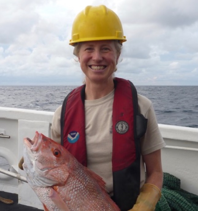 A smiling person wearing a yellow hard hat, red life vest, and beige shirt holds a large fish on a boat. The person is standing near the railing, with the ocean and cloudy sky in the background.