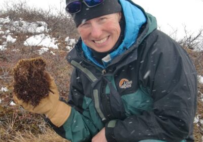 A person wearing outdoor winter gear, including a dark beanie, sunglasses, and gloves, smiles at the camera while holding a clump of moss or lichen. Snow is visible on the ground around them, indicating a cold, snowy environment.
