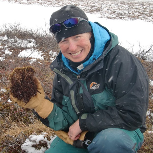 A person wearing outdoor winter gear, including a dark beanie, sunglasses, and gloves, smiles at the camera while holding a clump of moss or lichen. Snow is visible on the ground around them, indicating a cold, snowy environment.