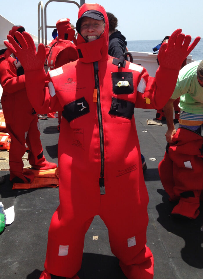 A person smiles and raises their hands while wearing a bright red survival suit on a ship with others donning similar suits in the background. The scene suggests a safety drill or training exercise at sea.