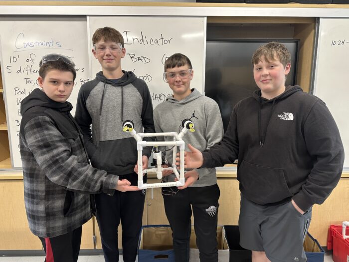 Four boys stand in a classroom holding a white PVC pipe cube structure. They wear safety goggles and casual clothing, and they are in front of a whiteboard with brainstorming notes. One wears a checkered jacket while the others wear hoodies.