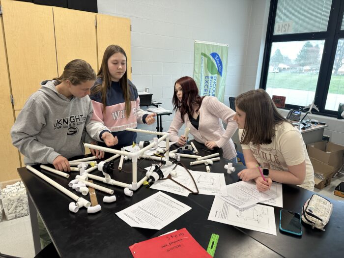 Four students collaborate on a project involving PVC pipes and connectors at a classroom table. They appear focused, using printed instructions and brainstorming together. A banner in the background reads "STEM" and some other equipment is visible around the room.