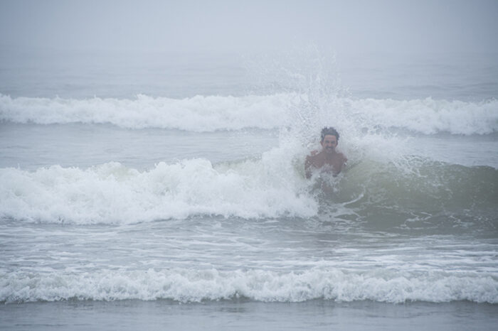 A person is partially submerged in ocean waves with a misty and foggy background. Water splashes around them as they face the camera, standing in the shallow water. The atmosphere appears overcast and serene.