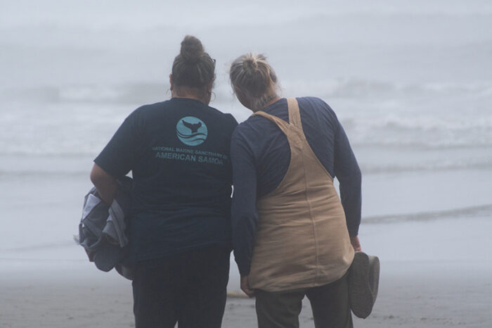 Two people stand closely together on a foggy beach, looking out at the misty ocean waves. One wears a dark T-shirt and holds a jacket, while the other wears a beige apron over a long-sleeve shirt and holds a hat. The ocean is blurred in the background.