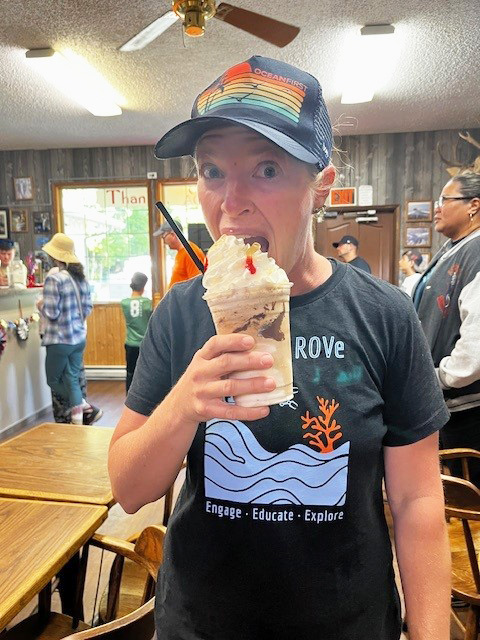 A person wearing a cap and a black t-shirt with graphics is eagerly about to sip a tall glass of a whipped cream-topped beverage with a cherry on top, inside a rustic café. Other patrons and wooden tables are visible in the background.