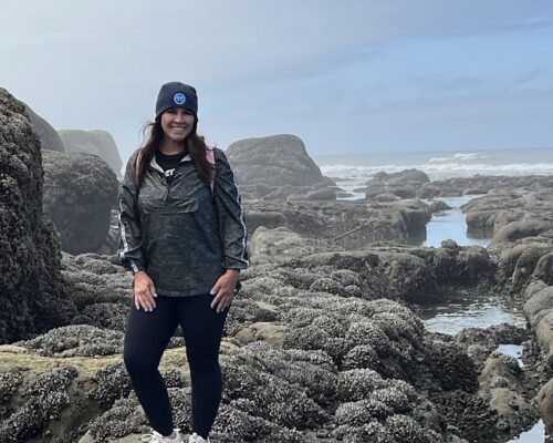 A person stands on a rocky coastal terrain with tide pools and large rocks around them. They wear a black beanie, grey jacket, black leggings, and white sneakers, with a pink backpack. The sky is cloudy, and waves can be seen in the background.