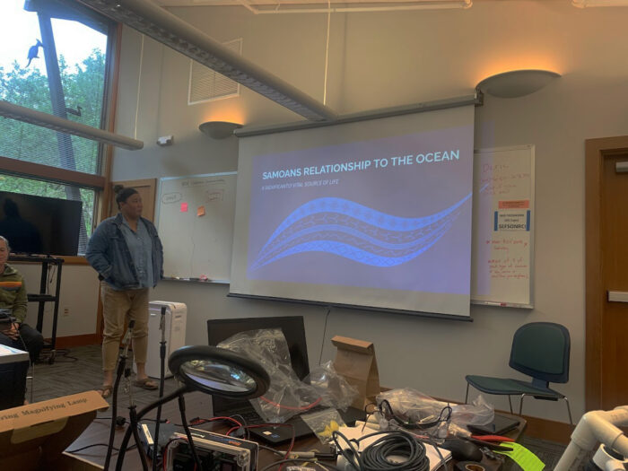 A person stands next to a projector screen with a presentation titled "Samoans Relationship to the Ocean" with a subtitle "A Foundation to Our Way of Life." The room contains various equipment, desks, and people, creating a classroom or workshop atmosphere.