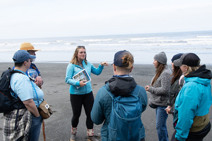 A group of people stands on a beach listening to a woman speaking to them. The speaker, holding a booklet, gestures with her left hand. The group is dressed in casual outdoor attire, and the ocean waves are visible in the background.