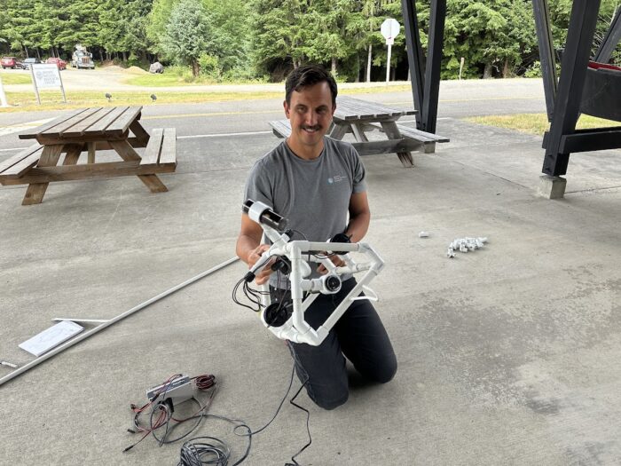 A person with short hair and a mustache is kneeling on a concrete floor under a shelter, assembling a white, geometric frame with various wires and components. There are picnic tables, a road, and trees in the background.