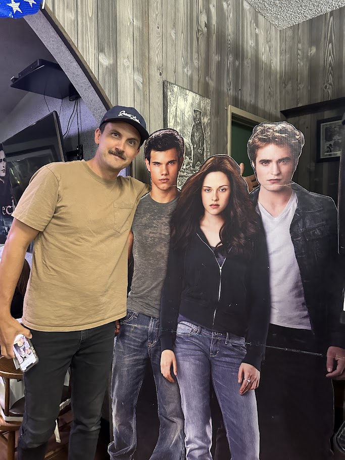 A man stands smiling next to cardboard cutouts of three characters from the Twilight series. The background features wood-paneled walls and various pictures and decorations. The man wears a hat and holds his phone in one hand.